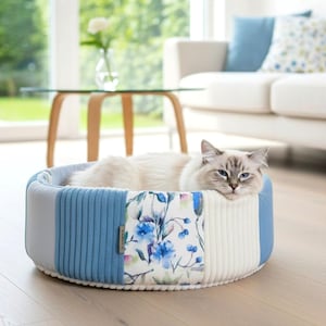May include: A round cat bed with a blue and white color scheme. The bed features a corduroy texture and a floral pattern. A fluffy cat is resting inside the bed.