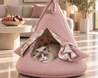 May include: A dusty pink felt pet tent with a triangular shape and a pointed top. The front opening features tie-back curtains with a floral pattern in shades of pink and green. A small rectangular tag labeled PRASKA is attached to the top.