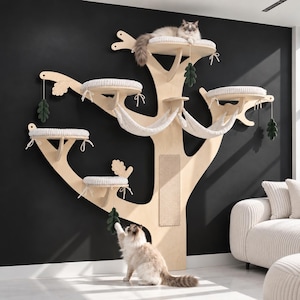 May include: A wall-mounted cat tree made of light-colored wood, featuring multiple circular platforms with white cushions, two fabric hammocks, a vertical scratching panel, and hanging dark green felt leaf toys, with two cats interacting with the structure.