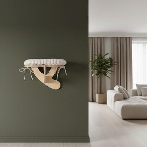 May include: A wall-mounted cat bed with a light wood frame and a cream-colored, cushioned top. The bed is secured with white ties and is designed to provide a comfortable resting spot for a cat. The bed is mounted on a dark green wall.