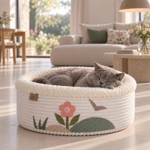 Puede incluir: Una cama redonda para mascotas hecha de cuerda color crema con un borde de felpa texturizado. El exterior presenta una aplicación decorativa de una flor rosa, hojas verdes, una colina color canela, una colina verde y un pájaro marrón.