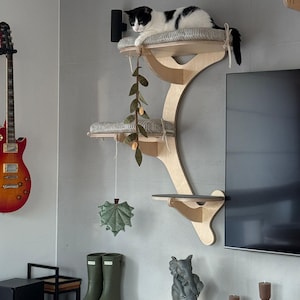 May include: A wall-mounted cat tree made of light-colored wood with three tiered platforms. The top two platforms feature soft, grey-striped cushions. A hanging felt leaf toy and a vine-like decoration dangle from the structure.