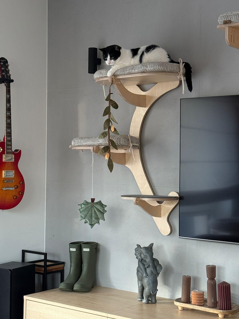 May include: A wall-mounted cat tree made of light-colored wood with three tiered platforms. The top two platforms feature soft, grey-striped cushions. A hanging felt leaf toy and a vine-like decoration dangle from the structure.