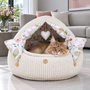 Puede incluir: Una cama para mascotas de pana color crema en forma de cúpula, con forro interior de estampado floral, un juguete blanco colgante en forma de corazón y botones de madera en el exterior. Un gato atigrado naranja descansa en su interior.