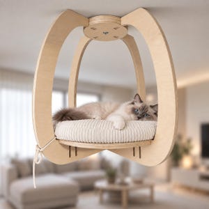 Könnte beinhalten: Ein helles Katzenbett aus Holz, das von der Decke hängt, mit einem geschwungenen Rahmen und einer runden Plattform. Ein flauschiges, cremefarbenes Kissen aus Kordstoff liegt auf der Basis, belegt von einer langhaarigen Katze mit blauen Augen.