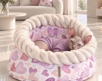 May include: A round pet bed with a thick, cream-colored plush braided rim and a white base featuring a purple and pink heart pattern. A hairless cat wearing a light purple shirt rests inside. A small rectangular tag labeled PRASKA is attached.