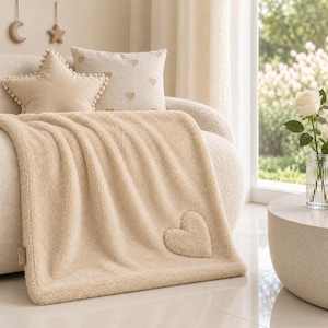Può includere: Una morbida coperta in pile color beige con un'applicazione a forma di cuore testurizzato nell'angolo inferiore. La coperta presenta un pelo spesso e soffice con un bordo rifinito, drappeggiata su un divano chiaro con cuscini abbinati.