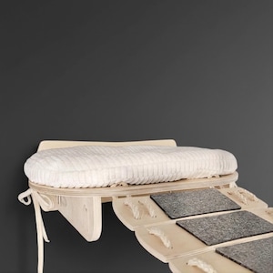 May include: A wooden cat shelf with a cream-colored, textured cushion and several gray, textured stepping stones. The shelf is designed to be mounted on a wall, with a rope tie on the side.