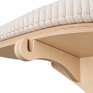 May include: A wall-mounted shelf featuring a light-colored plywood bracket with a curved support arm and a circular platform. The top of the platform is covered with a thick, cream-colored, ribbed fabric cushion that has a soft, textured surface.