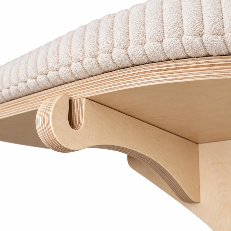 May include: A wall-mounted shelf featuring a light-colored plywood bracket with a curved support arm and a circular platform. The top of the platform is covered with a thick, cream-colored, ribbed fabric cushion that has a soft, textured surface.