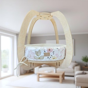 May include: A hanging cat bed with a light wood frame and a floral patterned interior. The bed is suspended from the ceiling and features a soft, cushioned interior with a cream-colored trim. The floral pattern includes pink and yellow flowers.