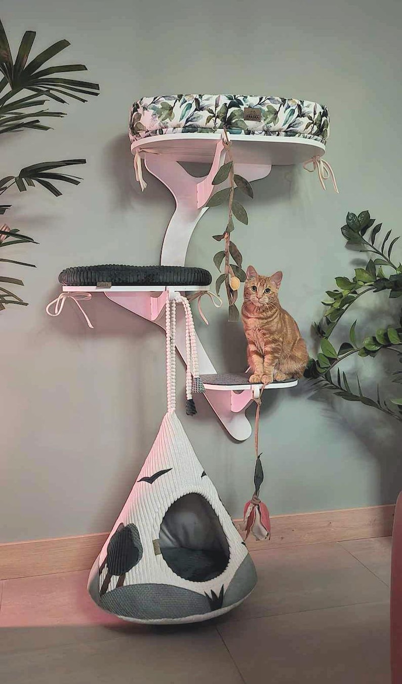 May include: A white wall-mounted cat tree with multiple levels, featuring a top platform with a botanical print cushion, a middle platform with a dark gray cushion, and a hanging white cone-shaped fabric cat bed with black bird graphics.