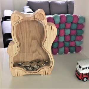 Op de afbeelding: Houten spaarpot in de vorm van een kat met een transparant plastic venster dat munten laat zien. De spaarpot staat op een wit oppervlak naast een roze en turquoise viltballenmat en een rode speelgoedbus.
