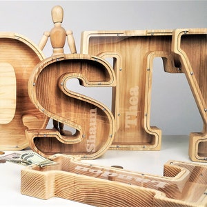 Può includere: Salvadanaio di legno a forma delle lettere "OSY" con un fronte in plastica trasparente. Le lettere sono su una superficie bianca. Le lettere hanno i nomi "Quinn", "Shaun" e "Thea" scritti su di esse.