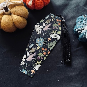 May include: A black coffin-shaped bookmark with a floral and botanical design. The bookmark features a hand reaching out from the flowers, a skull, and crystals. The bookmark has a black tassel.