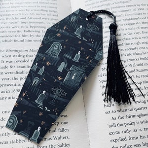 May include: A black bookmark with a spooky graveyard design featuring tombstones, skulls, and trees. The bookmark has a black tassel.