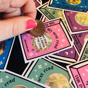 May include: A collection of rectangular scratch-off stickers featuring the text My next read and TBR INSPIRATION. Each sticker has a central holographic circle and celestial motifs. One sticker is being scratched with a coin to reveal hidden text.