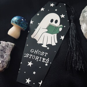 May include: Black and white bookmark shaped like a coffin with a ghost reading a book. The bookmark has white stars and the text "GHOST STORIES" on it. It has a black tassel.