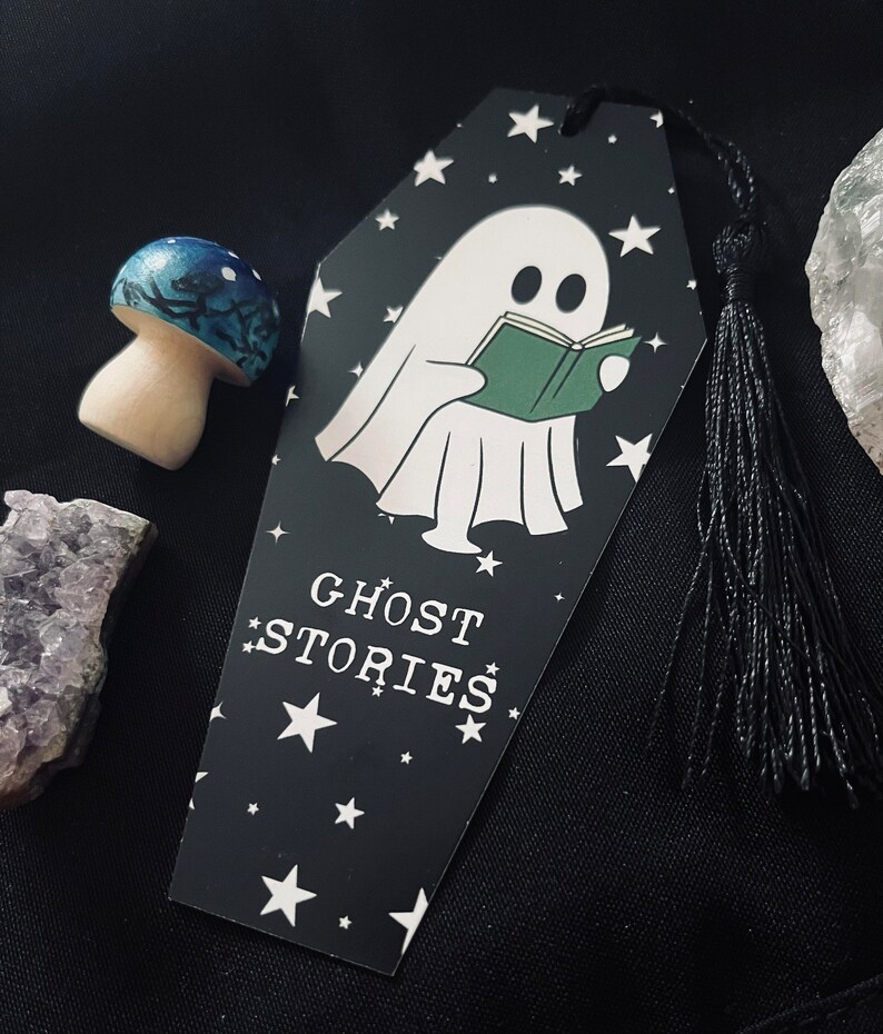 May include: Black and white bookmark shaped like a coffin with a ghost reading a book. The bookmark has white stars and the text "GHOST STORIES" on it. It has a black tassel.