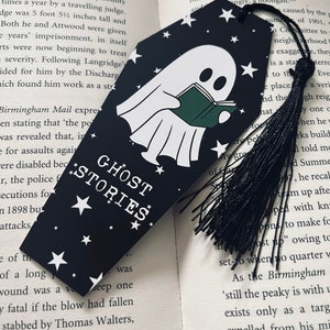 May include: A black bookmark with a white ghost reading a green book. The bookmark has white stars and the text "Ghost Stories" in white letters. A black tassel hangs from the bottom of the bookmark.