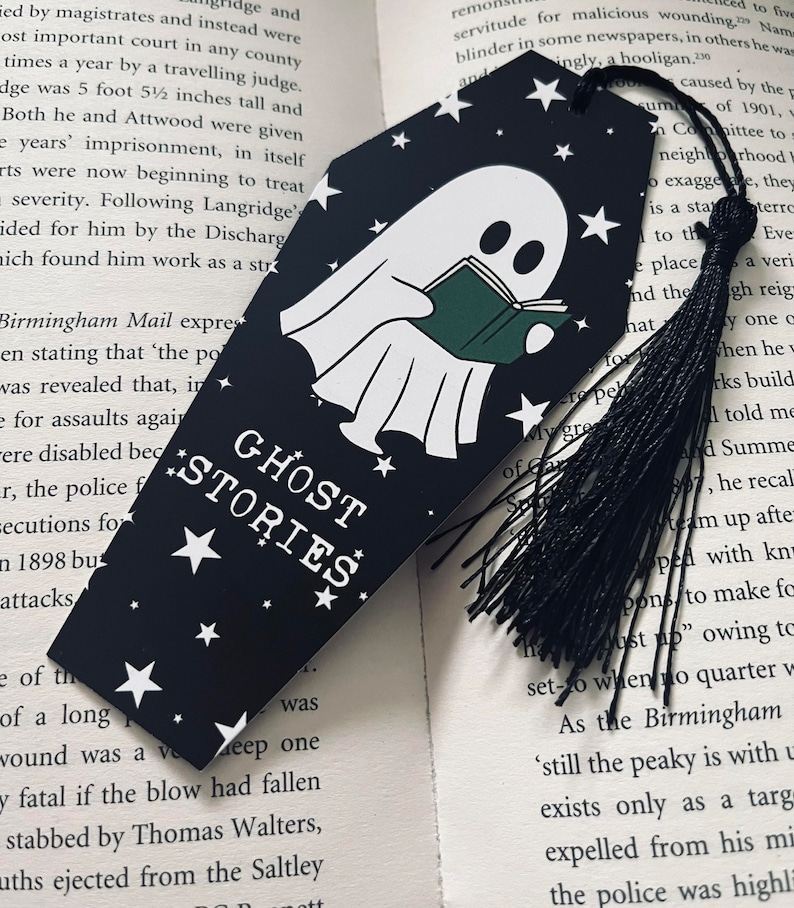 May include: A black bookmark with a white ghost reading a green book. The bookmark has white stars and the text "Ghost Stories" in white letters. A black tassel hangs from the bottom of the bookmark.