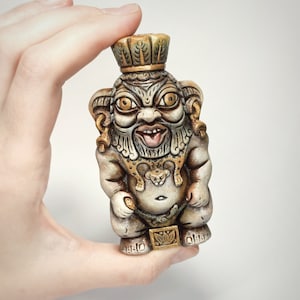 May include: A ceramic figurine of a mythical creature with a crown, wearing a gold necklace with a lion head pendant. The creature has a large, expressive face with a wide mouth and large eyes.