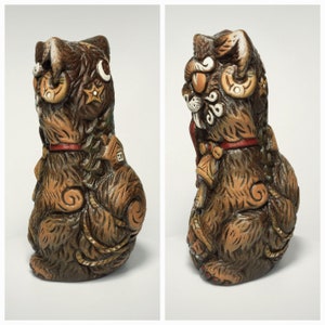 May include: A brown ceramic cat figurine featuring textured fur, a red collar, and decorative embossed symbols including a crescent moon, stars, and a small house. The cat has a stylized appearance with swirling patterns on its body and tail.