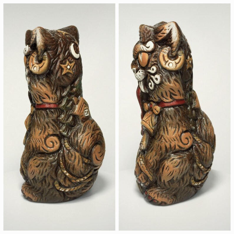 May include: A brown ceramic cat figurine featuring textured fur, a red collar, and decorative embossed symbols including a crescent moon, stars, and a small house. The cat has a stylized appearance with swirling patterns on its body and tail.