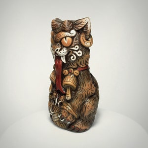 May include: A brown ceramic tiki mug shaped like a stylized feline creature with large orange eyes, a long red tongue, and white fangs. The figure features intricate carved details, including swirling patterns on the face and two golden bells hanging from a red collar around its neck.