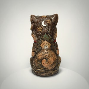 May include: A brown, sculpted figurine of a cat viewed from the back. The cat's fur is textured with carved details including a small house, a crescent moon, and stars. The figure features a curled tail and a red collar accent near the neck area.