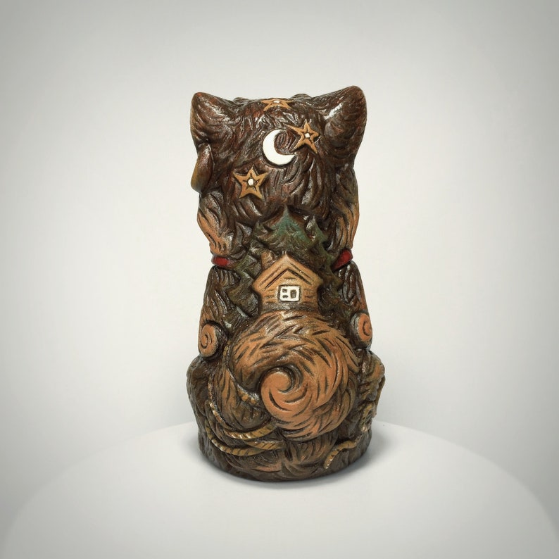 May include: A brown, sculpted figurine of a cat viewed from the back. The cat's fur is textured with carved details including a small house, a crescent moon, and stars. The figure features a curled tail and a red collar accent near the neck area.