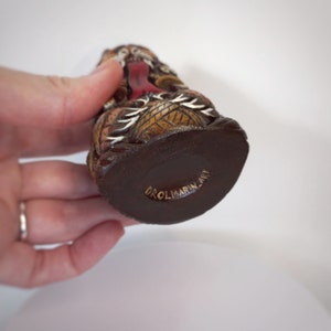 May include: A small, hand-carved wooden figurine featuring intricate brown, black, and white details with a prominent red tongue. The base of the object is dark brown and features the gold-colored text DROLMARIN_ART engraved on the bottom surface.