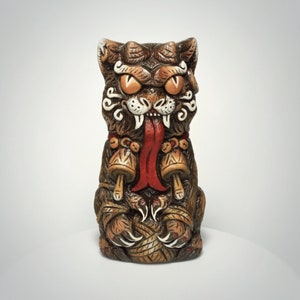 May include: A brown ceramic tiki mug shaped like a stylized cat with large orange eyes, a long red tongue, and white fangs. The cat figure features intricate carved details, including a collar with two bell-like ornaments and sharp claws.