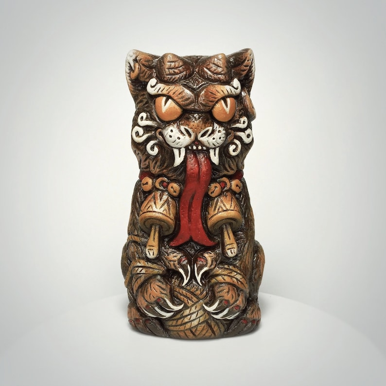 May include: A brown ceramic tiki mug shaped like a stylized cat with large orange eyes, a long red tongue, and white fangs. The cat figure features intricate carved details, including a collar with two bell-like ornaments and sharp claws.