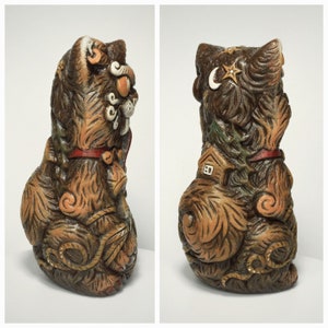 May include: A brown ceramic cat figurine featuring intricate carved details including a crescent moon, stars, a small house, and pine trees. The cat wears a red collar with a bell and has textured fur patterns throughout its entire body surface.