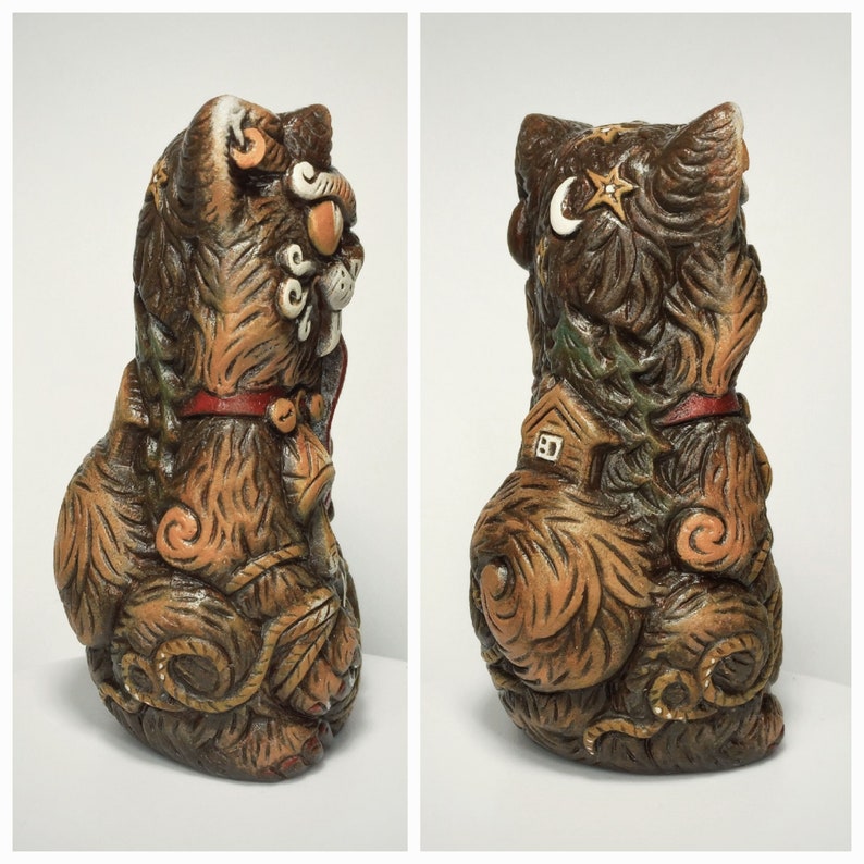 May include: A brown ceramic cat figurine featuring intricate carved details including a crescent moon, stars, a small house, and pine trees. The cat wears a red collar with a bell and has textured fur patterns throughout its entire body surface.