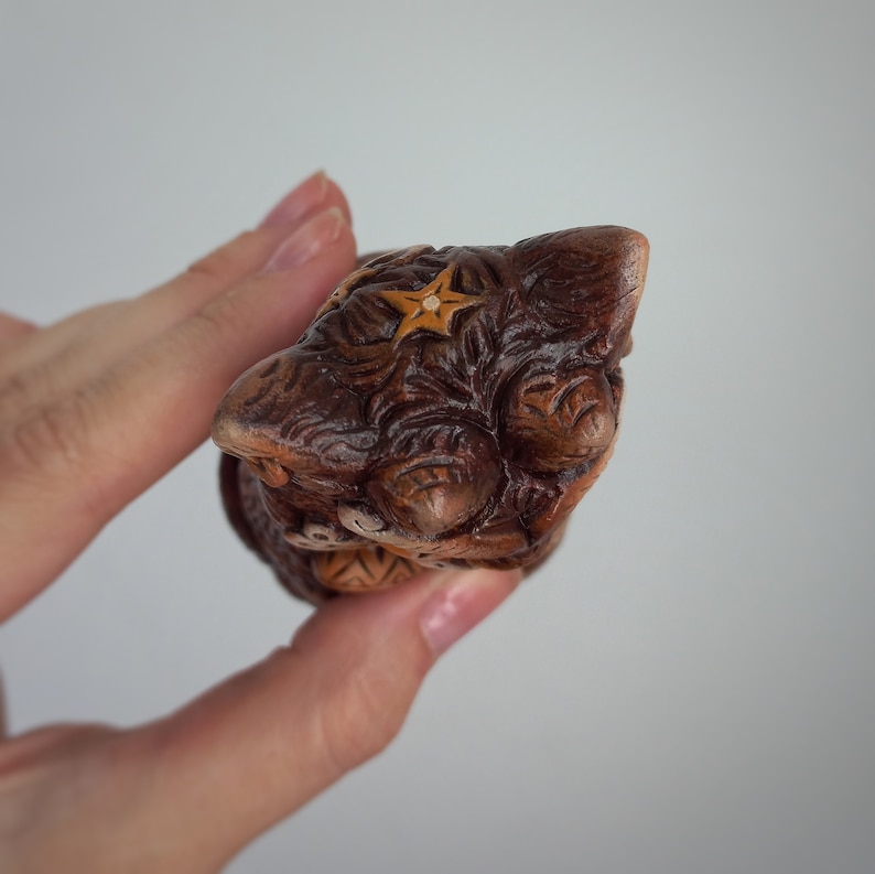 May include: A small, hand-carved wooden figurine of a cat with intricate, swirling patterns etched into the dark brown wood. The top of the head features a small, light-colored star carving, and the surface displays a polished, textured finish.