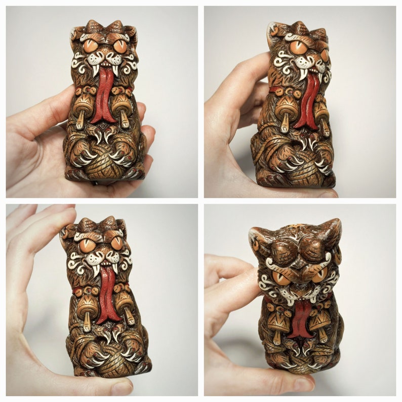 May include: A brown, cat-like creature figurine with orange eyes, a long red tongue, and white fangs. The creature has textured fur, wears a collar with two small mushroom-shaped charms, and holds a ball of yarn between its front paws.