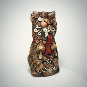 May include: A brown, sculpted cat figurine with a fierce expression, featuring large orange eyes, a long red tongue, and sharp white fangs. The cat wears a collar with two bell-shaped charms and holds a ball of yarn with its clawed paws.