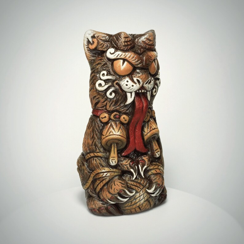 May include: A brown, sculpted cat figurine with a fierce expression, featuring large orange eyes, a long red tongue, and sharp white fangs. The cat wears a collar with two bell-shaped charms and holds a ball of yarn with its clawed paws.