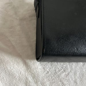 May include: A black rectangular handbag with a shoulder strap. The bag is made of a smooth, possibly leather material. The strap is thin and appears to be adjustable. The bag is closed with a flap.