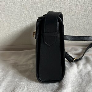 May include: Black leather crossbody bag with a rectangular shape and a flap closure. The bag features a gold-colored clasp and a long, adjustable strap. The bag is sitting on a white, textured surface.