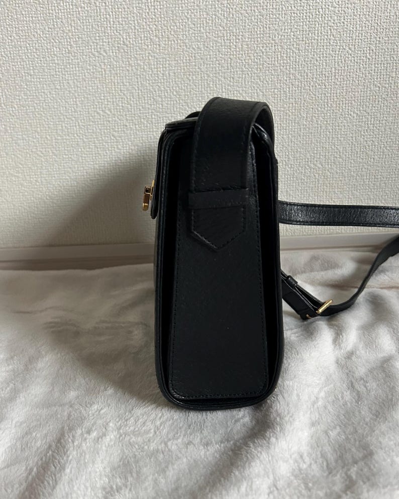 May include: Black leather crossbody bag with a rectangular shape and a flap closure. The bag features a gold-colored clasp and a long, adjustable strap. The bag is sitting on a white, textured surface.
