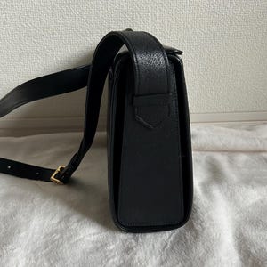 May include: Black leather crossbody bag with a long adjustable strap and a gold-colored buckle. The bag has a rectangular shape with a flap closure. The leather has a textured appearance.