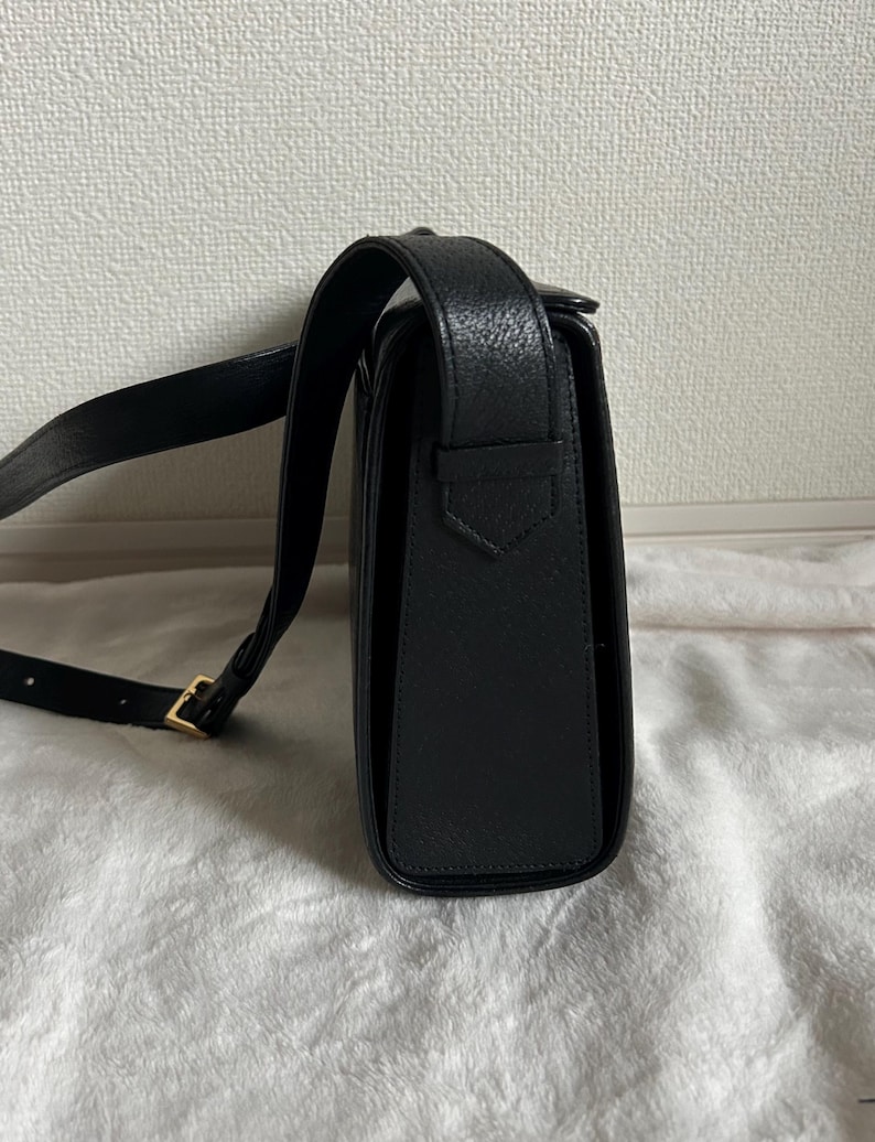 May include: Black leather crossbody bag with a long adjustable strap and a gold-colored buckle. The bag has a rectangular shape with a flap closure. The leather has a textured appearance.
