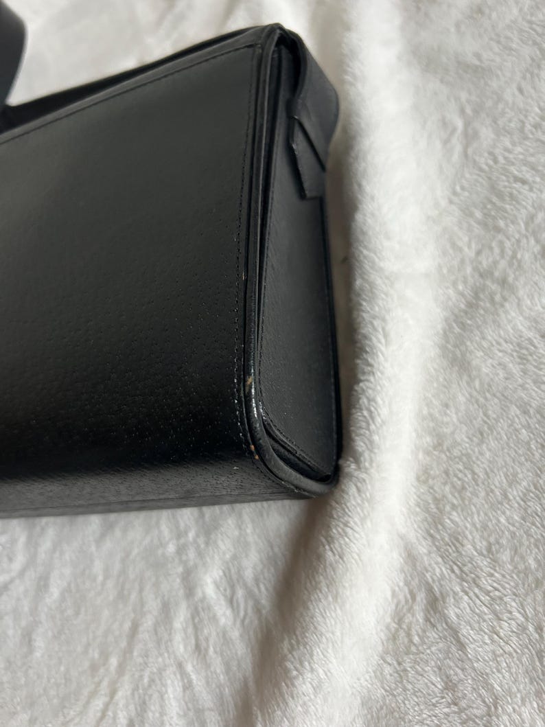 May include: A black rectangular handbag with a structured design. The bag has a smooth, matte finish and a visible seam detail along the edges. The bag's corner is slightly rounded.