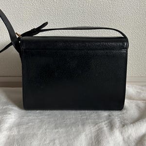 May include: Black rectangular crossbody bag with a textured surface. The bag has a long, adjustable strap and a small handle on top. The bag is sitting on a white, textured surface.