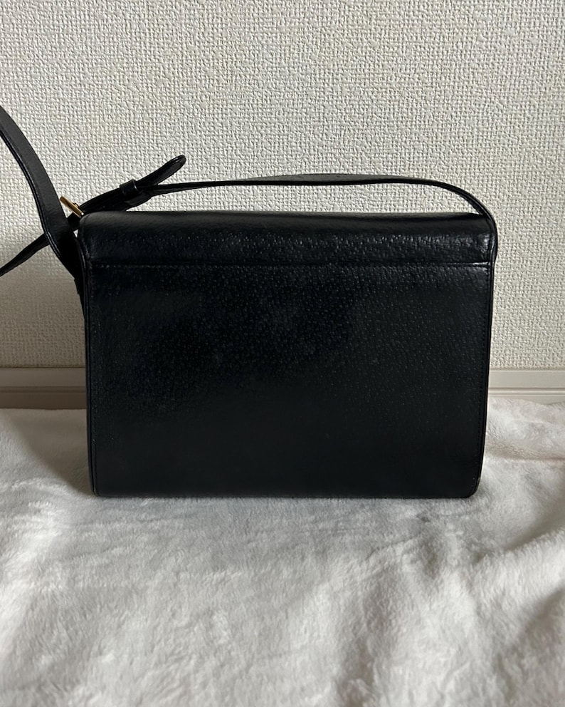 May include: Black rectangular crossbody bag with a textured surface. The bag has a long, adjustable strap and a small handle on top. The bag is sitting on a white, textured surface.