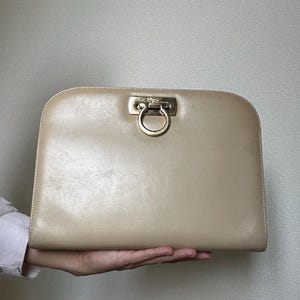 May include: A beige leather clutch bag with a rounded top edge and a gold-toned metal Gancini closure centered on the front. The smooth surface of the bag features subtle signs of wear, and the metallic hardware displays a polished finish.