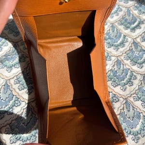 May include: Open, brown leather wallet with a gold-colored clasp. The wallet is open, revealing the interior compartments. The leather has a textured appearance. The wallet is on a patterned surface.